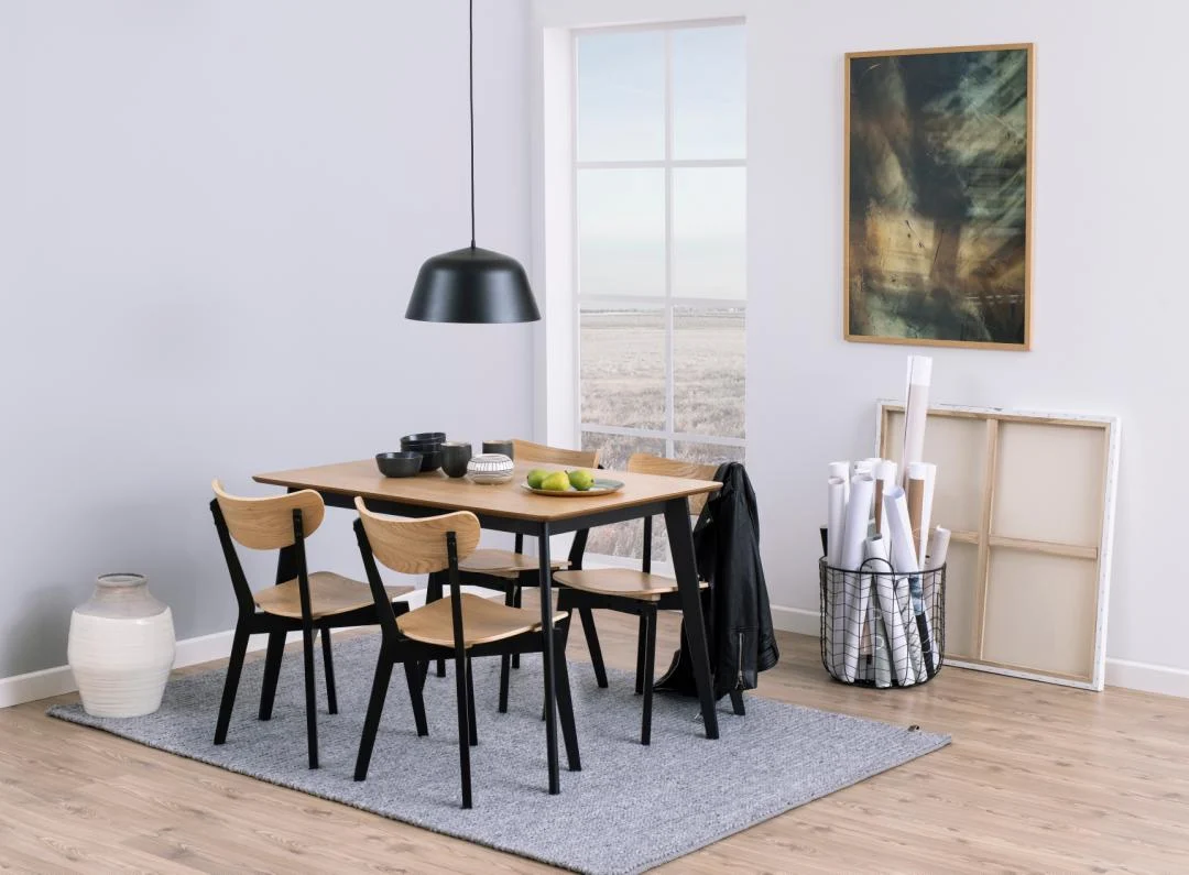 Een moderne eetkamer met de Eettafel Roxby 80 x 76 CM Rond Eikenhout met een matzwarte basis, vier stoelen, een zwarte hanglamp en een grijs vloerkleed. Een raam biedt uitzicht op buiten, terwijl opgerolde papieren, kunst en een grote vaas de hoek sieren.