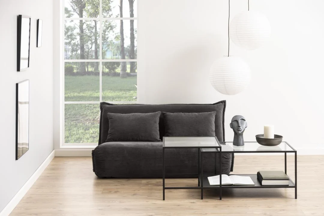 Een minimale woonkamer heeft een lage, donkergrijze bank en twee Salontafel Zwart 50 x 45 CM Rechthoek kunststof tafels met decor. Een groot raam, witte muren en hangende papieren lantaarns maken de lichte, moderne sfeer compleet.
