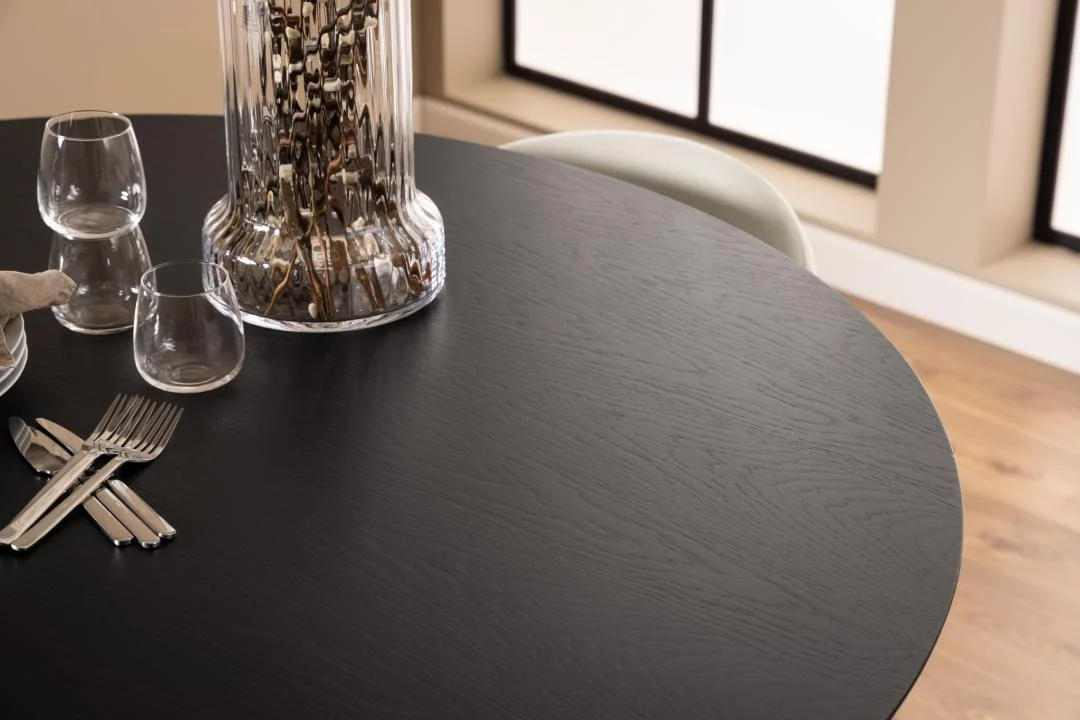 Een close-up van de Eettafel Nieuwe Sfeer 140 CM Rond in eikenhout, bezet met glaswerk, zilverwerk en een glazen vaas, staat naast een lichte stoel bij grote ramen.