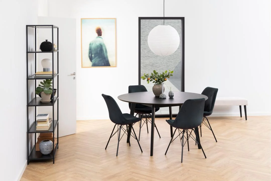Moderne eetkamer met de Eettafel Nieuwe Sfeer 140 CM Rond in eikenhout, vier zwarte stoelen, een plantaardig middenstuk, hanglamp met papieren lantaarn, metalen plank met decor, witte bank, minimalistische kunst op witte muren en houten vloer.