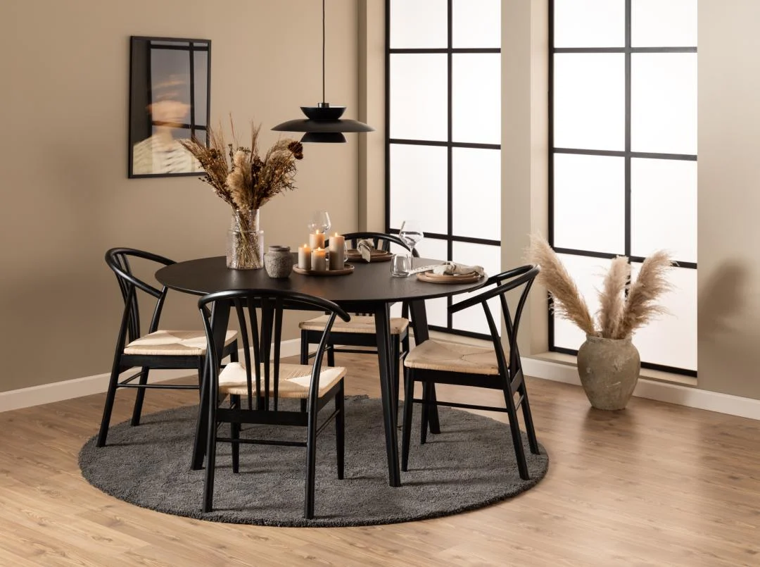 Een moderne eetkamer met de Eettafel Nieuwe Sfeer 140 CM Rond in eikenhout, vier zwarte geweven stoelen, kaarsen, droogbloemen en servies. Grote ramen, pampagras en een neutrale inrichting creëren een gezellige, minimalistische sfeer.