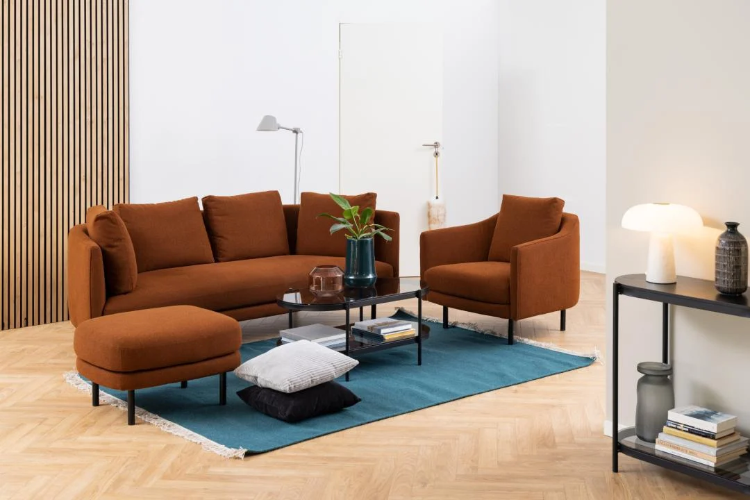 In een moderne woonkamer staan een roestkleurige bank, fauteuil en voetenbank op een blauw vloerkleed. De Salontafel Zwart, Grijs 50 x 42 CM Rond (kunststof) met boeken en een plant staat ervoor. Een consoletafel bevat decor en een lamp; de houten vloer maakt de look compleet.