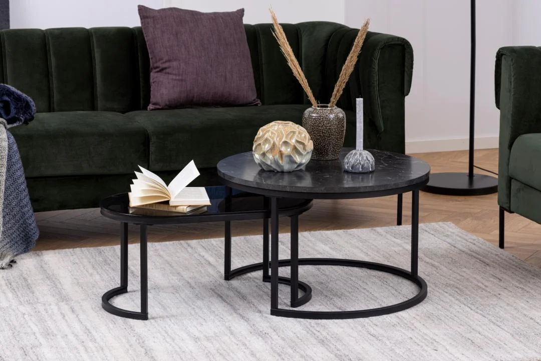 De Salontafel Rookglas, Zwart Rond Kunststof staat op een licht vloerkleed met vazen en een open boek, terwijl een groene bank met een paars kussen kleur toevoegt op de achtergrond. De setting is modern en gezellig.