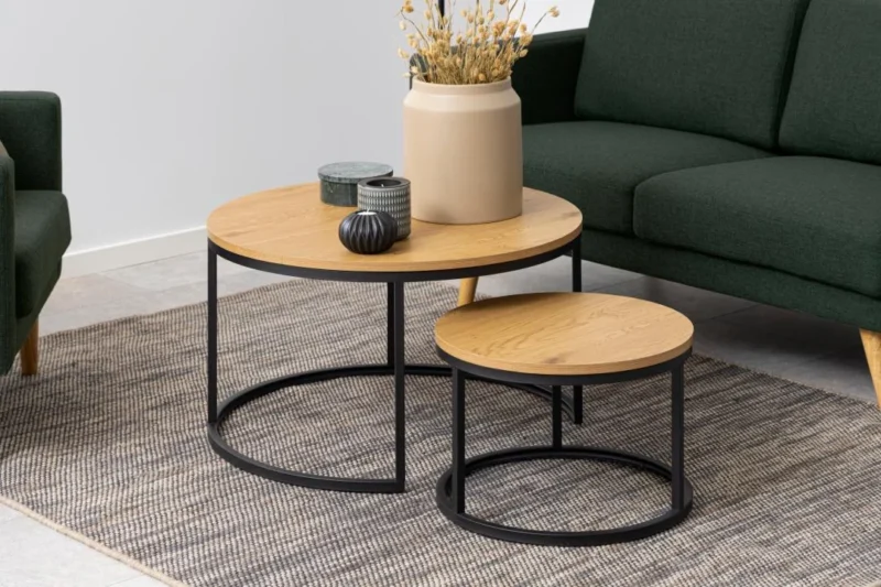 De Salontafel Rond Kunststof heeft twee ronde salontafels met zwarte metalen poten op een vloerkleed met structuur. Op de grootste tafel staan een beige vaas met gedroogde bloemen en twee decoratieve potten, terwijl groene banken gedeeltelijk zichtbaar zijn op de achtergrond.
