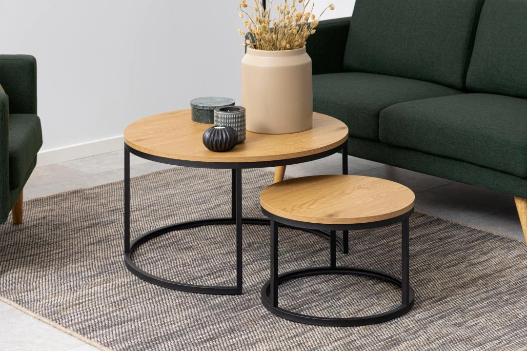 De Salontafel Rond Kunststof heeft twee ronde salontafels met zwarte metalen poten op een vloerkleed met structuur. Op de grootste tafel staan een beige vaas met gedroogde bloemen en twee decoratieve potten, terwijl groene banken gedeeltelijk zichtbaar zijn op de achtergrond.