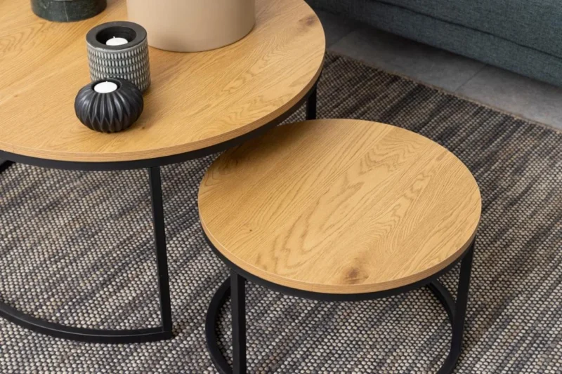 Twee Salontafel Rond Kunststof tafels met een zwart metalen onderstel staan op een geweven vloerkleed. Op de grotere tafel staan twee vazen en een kaars, terwijl de kleinere leeg is. Een grijze bank is gedeeltelijk zichtbaar op de achtergrond.