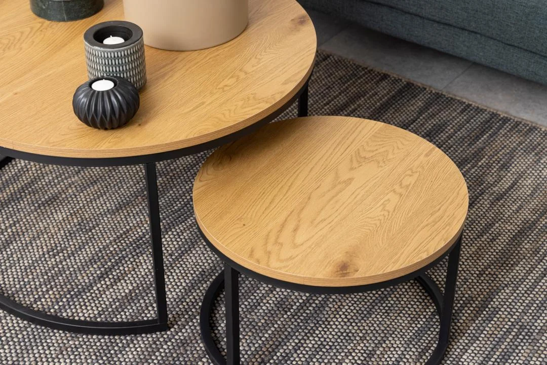 Twee Salontafel Rond Kunststof tafels met een zwart metalen onderstel staan op een geweven vloerkleed. Op de grotere tafel staan twee vazen en een kaars, terwijl de kleinere leeg is. Een grijze bank is gedeeltelijk zichtbaar op de achtergrond.