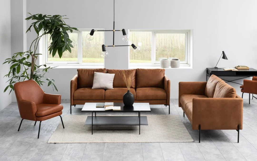 Moderne woonkamer met bruine leren banken en fauteuil, een Salontafel Zwart 60 x 40 CM Rechthoek in industrieel zwart, boeken en een vaas, grote potplant, bureau in de hoek, minimalistische inrichting en natuurlijk licht van twee ramen.