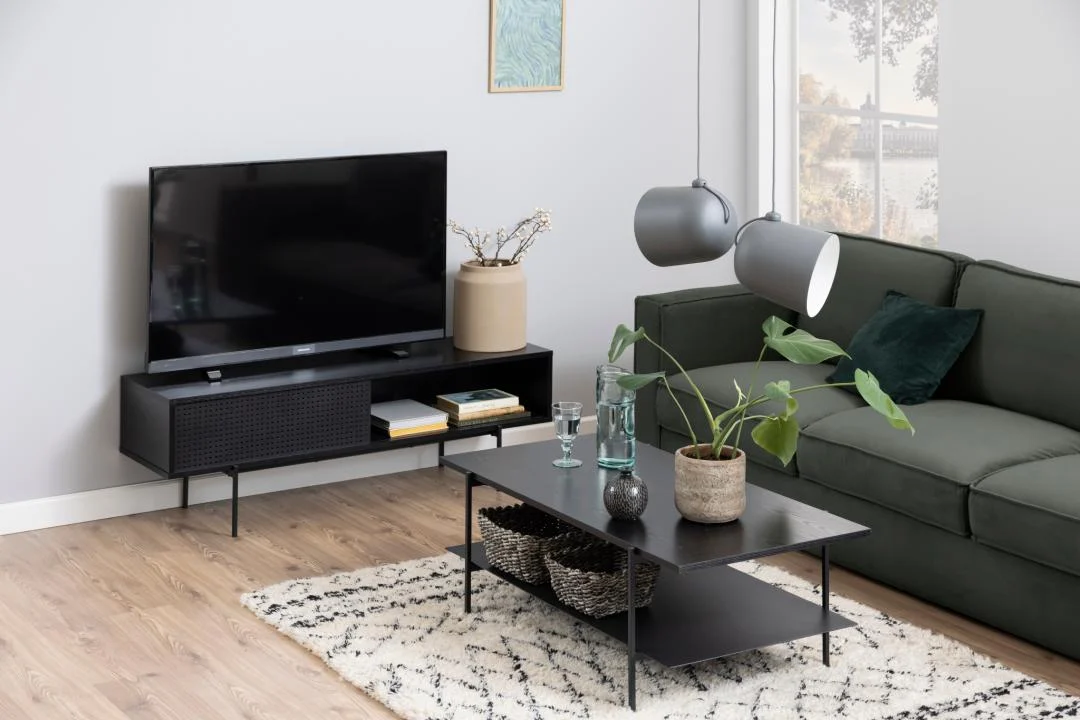 Een moderne woonkamer met een groene bank, Salontafel Zwart 60 x 40 CM Rechthoek (kunststof industrieel) met planten en glazen, een tv op een zwarte standaard, beige en wit vloerkleed, hanglampen en kunstwerken op een lichtgrijze muur.