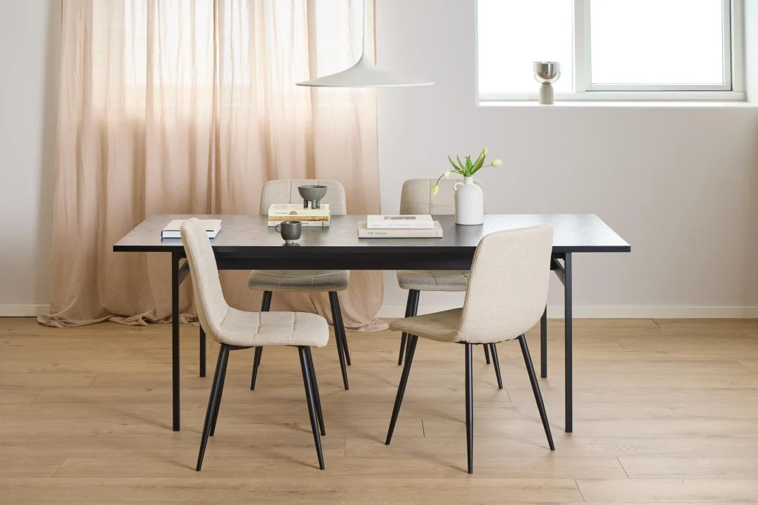 Een lichte, minimalistische eetkamer met de Eettafel Nieuwe Sfeer 90 x 74 CM rechthoek MDF zwarte tafel, vier beige stoelen, boeken, een vaas met witte bloemen, een kopje, een schaal, lichthouten vloeren, transparante gordijnen en een zonovergoten raam.