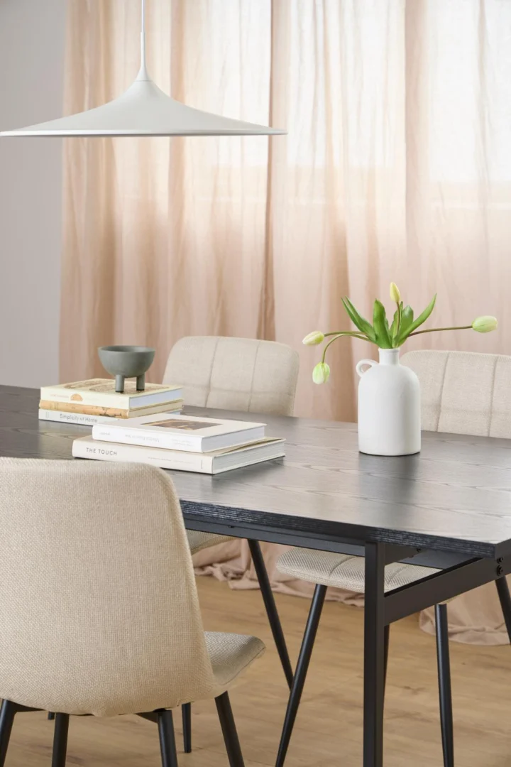 De Eettafel Nieuwe Sfeer (90 x 74 CM) met een rechthoekig MDF blad is vormgegeven met beige stoelen, boeken, een schaal en een witte tulpenvaas. Zachtroze gordijnen en een witte hanglamp maken deze lichte, minimalistische eetkamer compleet.