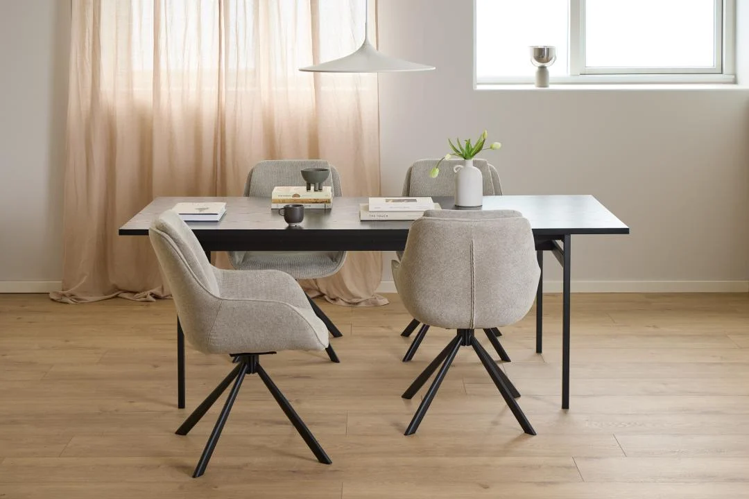 In een moderne eetkamer staat de Eettafel Nieuwe Sfeer (90 x 74 cm), een rechthoekige zwarte MDF tafel met vier lichtgrijs gestoffeerde stoelen met zwart metalen poten, boeken, een vaas en een kleine plant. Beige gordijnen en een raam maken de achtergrond compleet.