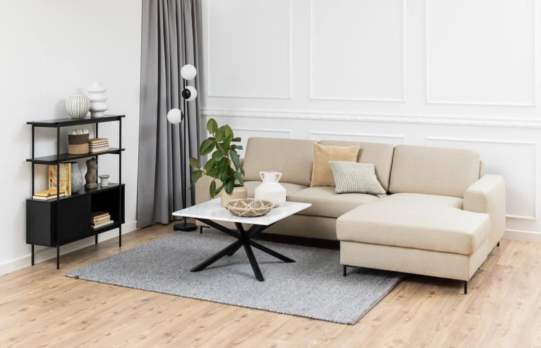 Moderne woonkamer met een beige sofa, witte marmeren salontafel, grijs vloerkleed, Angus boekenkast met decor, een plant in pot en grijze gordijnen tegen witte wanden met panelen op een houten vloer.
