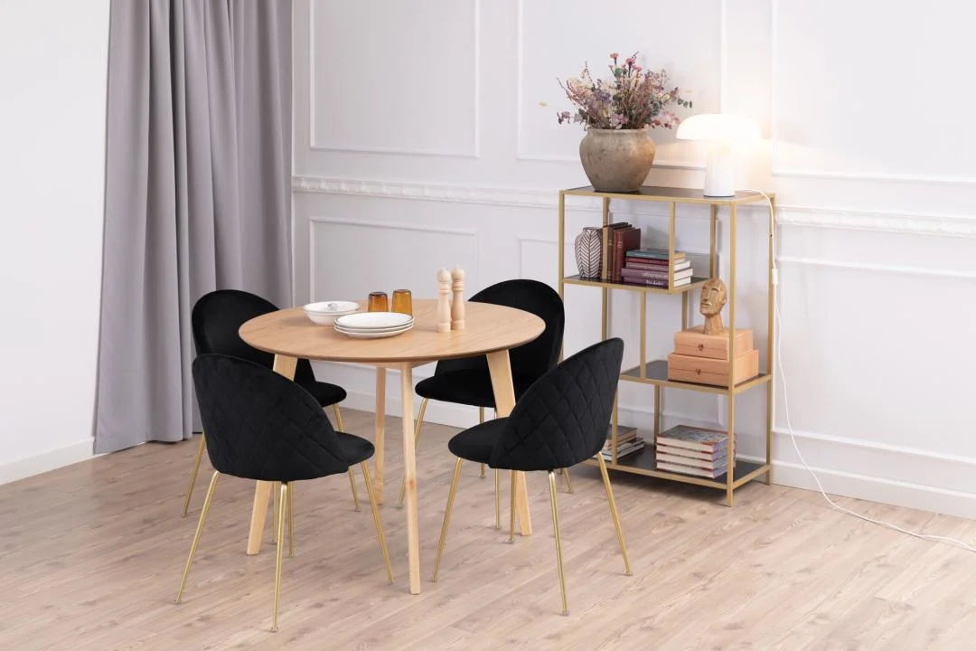 In een moderne eethoek staat de Eettafel Nieuwe Sfeer 105 CM Rond in massief eikenhout, vier zwarte stoelen met gouden poten en een goudkleurig rek met boeken, decor en een lamp. Neutrale muren, houten vloeren en grijze gordijnen versterken de minimalistische stijl.