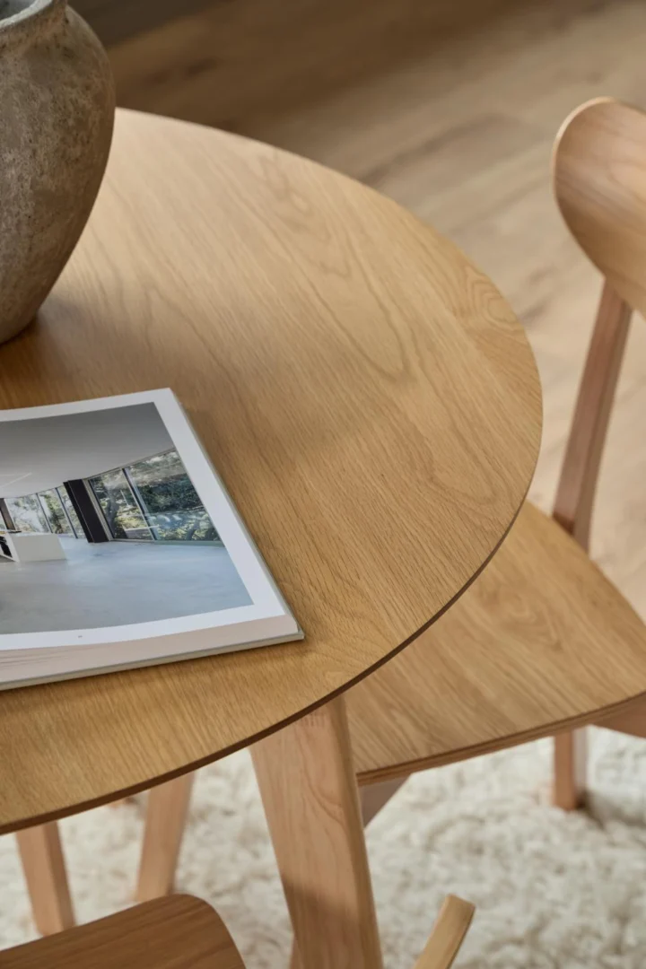 Een close-up van de Eettafel Nieuwe Sfeer 105 CM Rond in massief eiken met twee bijpassende stoelen. Een grote vaas en een open tijdschrift rusten op de tafel, die op een licht vloerkleed op een houten vloer ligt.