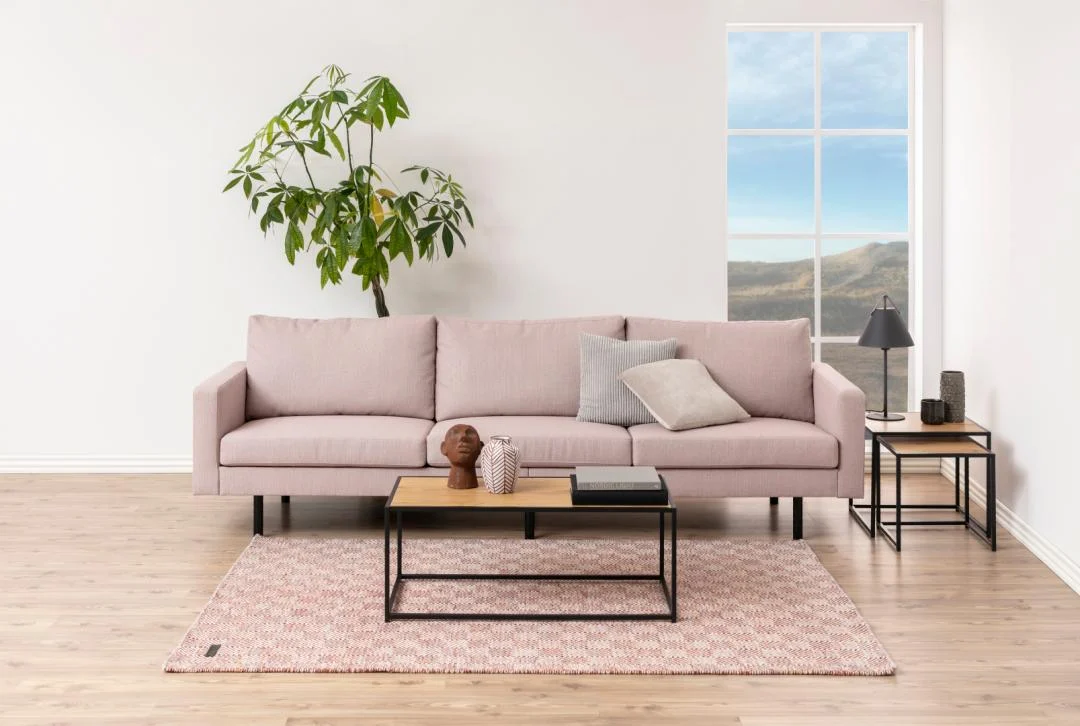 Een moderne woonkamer met een lichtroze bank, twee neutrale kussens, een Salontafel Zwart 50 x 40 CM Rechthoek van staal, een decoratieve plant, een bijzettafel met een lamp en een groot raam met een prachtig uitzicht op de buitenlucht.