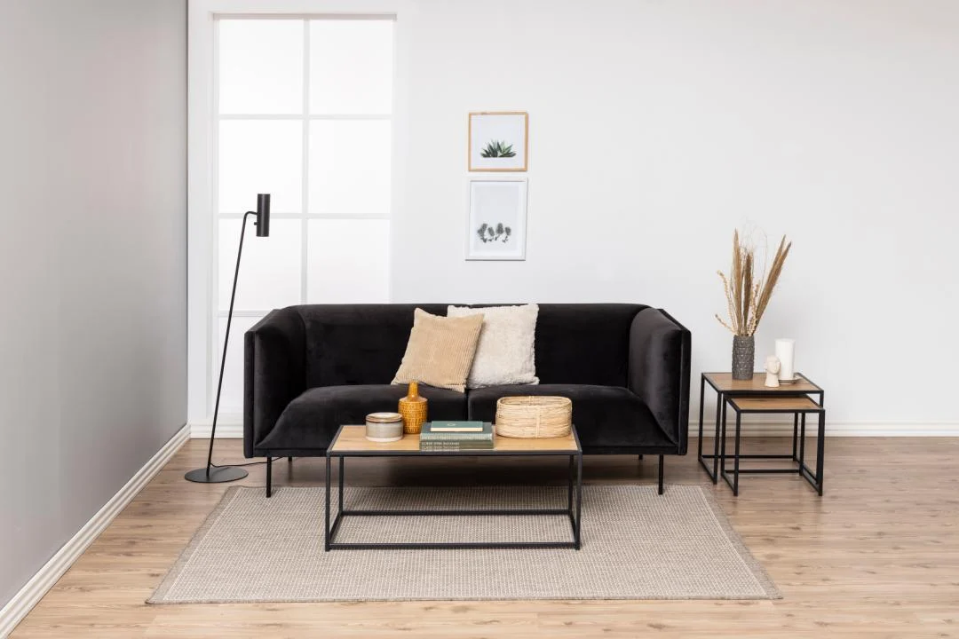 Een moderne woonkamer met een zwarte bank, beige kussens, staande lamp, minimalistische muurkunst en een rechthoekige Salontafel Zwart 50 x 40 CM (staal) met boeken en decor, plus bijzettafeltjes met vazen, op een licht vloerkleed op een houten vloer.
