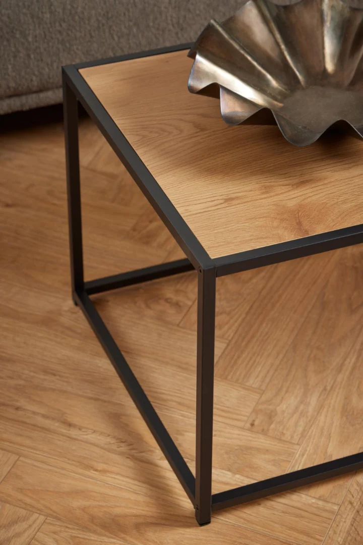 De Salontafel Zwart 50 x 40 CM Rechthoek van staal staat op een houten vloer, met een decoratieve golfrandige metalen schaal erop. Op de achtergrond is gedeeltelijk een grijze bank zichtbaar.