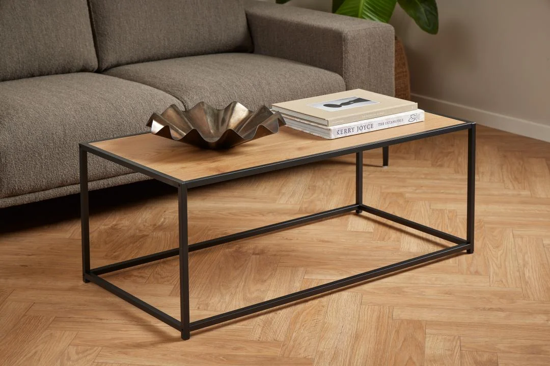 De Salontafel Zwart 50 x 40 CM Rechthoek, gemaakt van staal, staat op een vloer met visgraatmotief met een metalen middenstuk en tijdschriften erop. Op de achtergrond is een grijze bank zichtbaar.