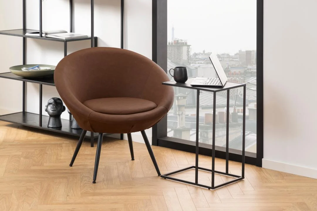 Een moderne kamer heeft een ronde bruine stoel, een Bijzettafel Zwart 35 CM Rechthoek met een laptop en mok, een boekenplank met decor en grote ramen met uitzicht op de stad.