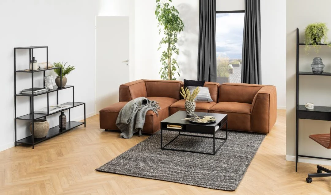 Moderne woonkamer met een bruine sectionele bank, de Salontafel Zwart 80 x 40 CM Vierkant met een kunststof opbergvak, grijs vloerkleed, open rekken met planten en decor, een grote groene plant en een raam met donkere gordijnen en natuurlijk licht.