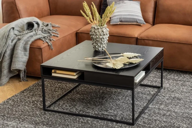 Een moderne woonkamer met een bruine leren bank, grijze plaids en een Salontafel Zwart 80 x 40 CM Vierkant met een kunststof opbergvak, een vaas met gedroogde planten, een zwart dienblad en boeken, alles op een grijs vloerkleed met structuur.