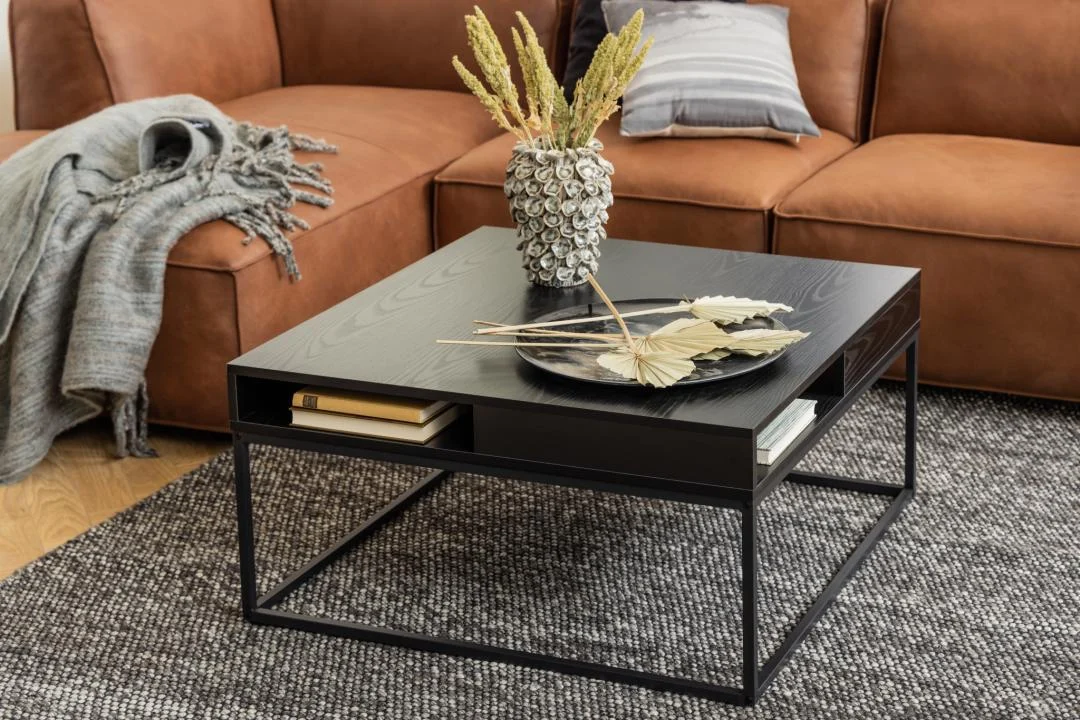 Een moderne woonkamer met een bruine leren bank, grijze plaids en een Salontafel Zwart 80 x 40 CM Vierkant met een kunststof opbergvak, een vaas met gedroogde planten, een zwart dienblad en boeken, alles op een grijs vloerkleed met structuur.