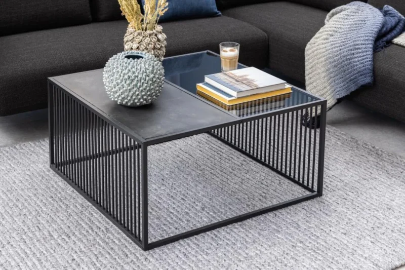 De Salontafel Rookglas, Zwart 80 x 40 CM met een vierkant blad van gerookt glas toont een vaas met textuur, gedroogde bloemen, een glas koffie en gestapelde boeken. Hij rust op een lichtgrijs vloerkleed naast een donkere bank gedrapeerd met een gebreide deken.