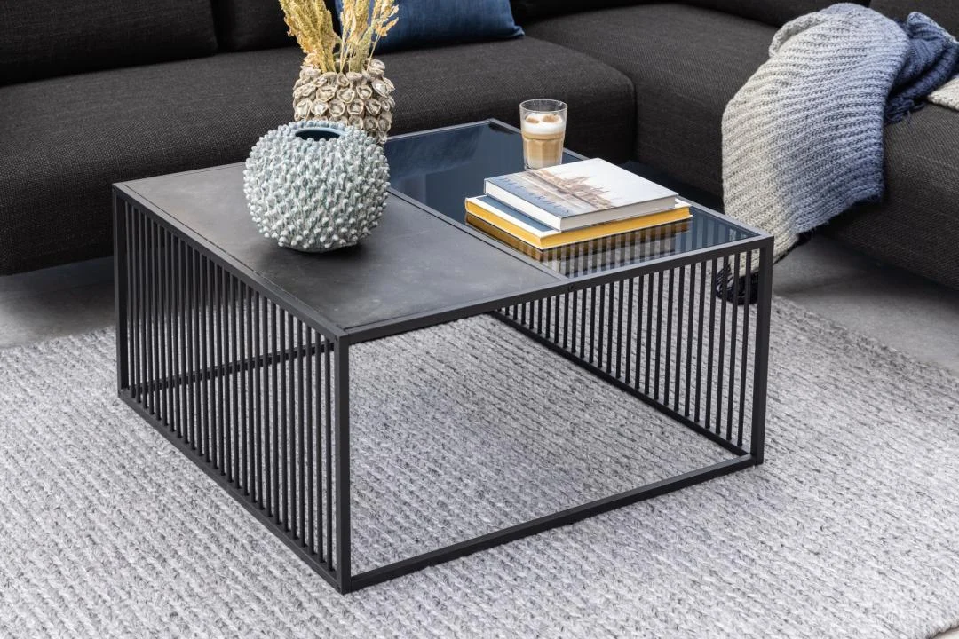 De Salontafel Rookglas, Zwart 80 x 40 CM met een vierkant blad van gerookt glas toont een vaas met textuur, gedroogde bloemen, een glas koffie en gestapelde boeken. Hij rust op een lichtgrijs vloerkleed naast een donkere bank gedrapeerd met een gebreide deken.