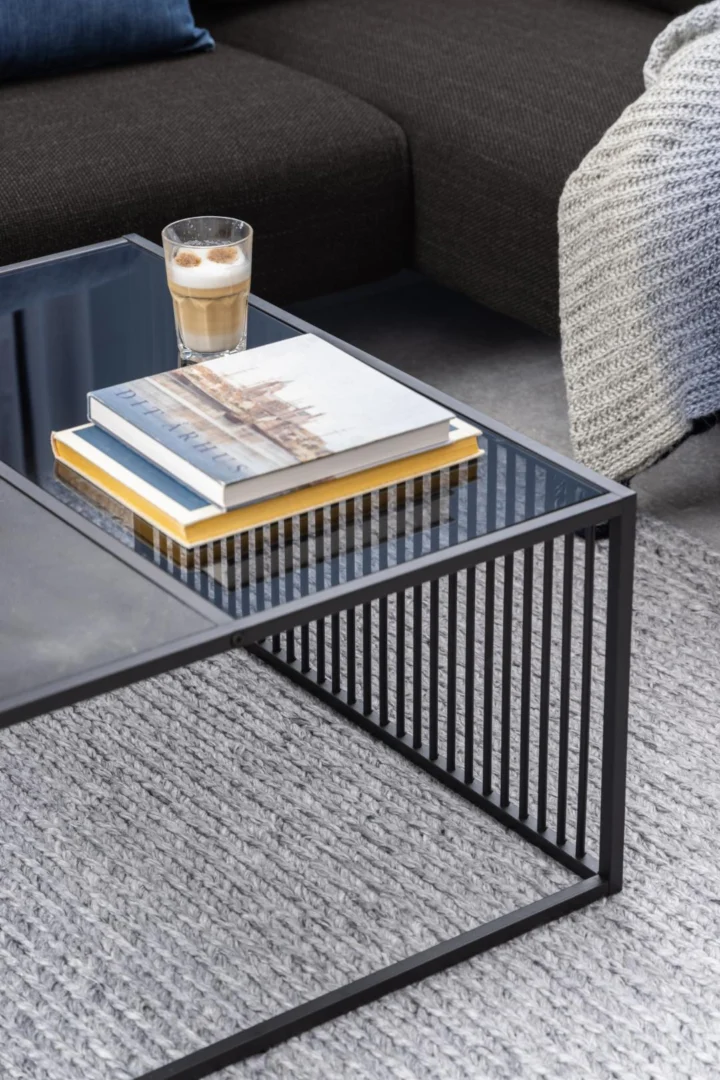Op een Salontafel Rookglas, Zwart 80 x 40 CM met vierkant rookglas en zwart frame staan een koffie, twee boeken en een tijdschrift. De tafel staat op een grijs vloerkleed met structuur bij een donkere bank gedrapeerd met een lichtgrijze gebreide deken.