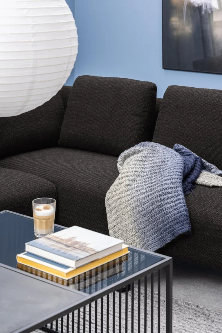 Een gezellige woonkamer met een donkere bank, grijze gebreide deken, gestapelde boeken en koffie op de Salontafel Rookglas, Zwart (80 x 40 cm) met vierkant rookglas, alles tegen lichtblauwe muren en een moderne inrichting.