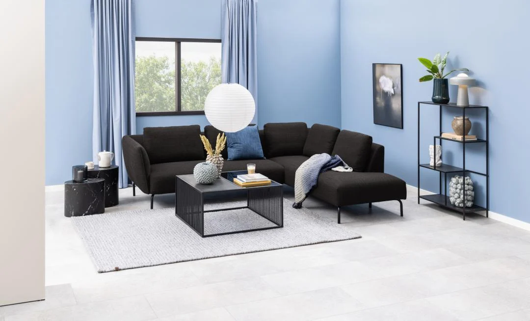 Een moderne woonkamer met een zwarte sectionele bank, vierkant rookglas Salontafel Rookglas, Zwart 80 x 40 CM, licht vloerkleed, blauwe muren, grote ramen met lichtblauwe gordijnen, een zwarte plank met decor, muurkunst en een ronde papieren lantaarnlamp.