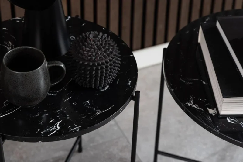 Twee Bijzettafels Zwart 52 CM Rond Kunststof: op de ene staat een donkere mok en een stekelig decor, op de andere een stapel boeken. De vloer is betegeld, met een houten lattenwand op de achtergrond.