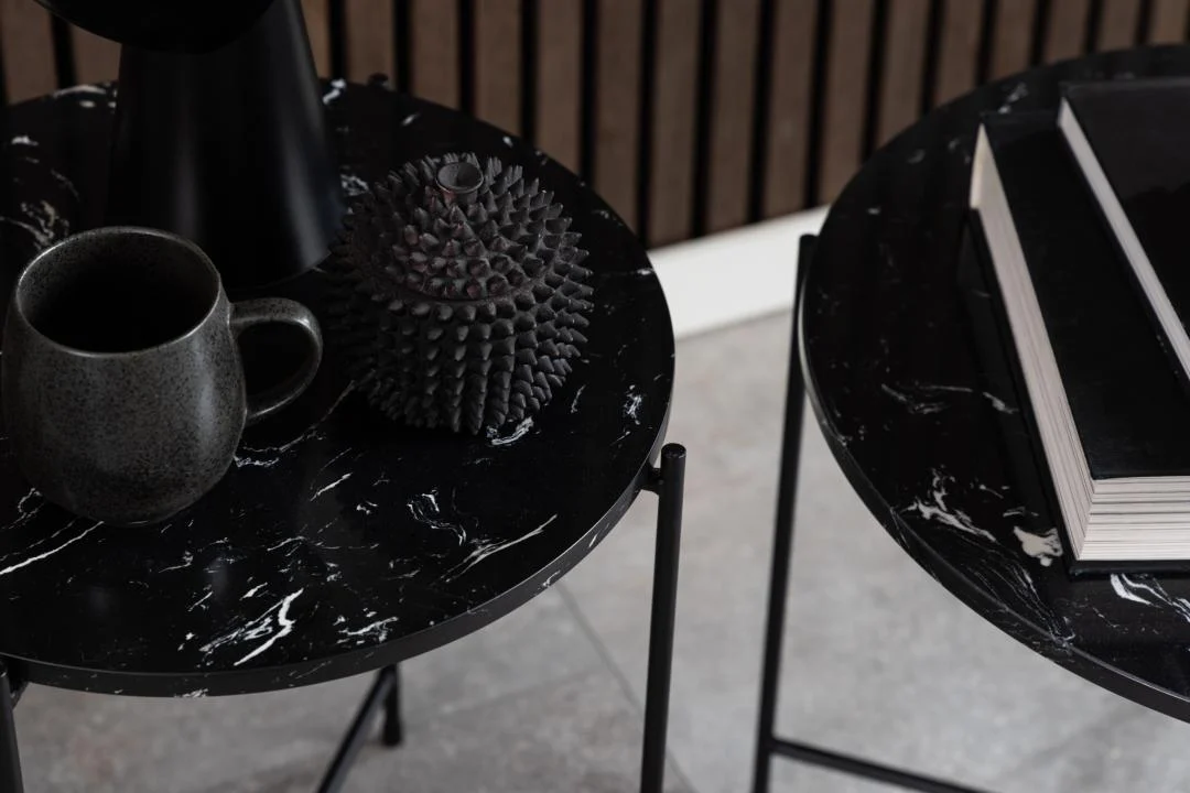 Twee Bijzettafels Zwart 52 CM Rond Kunststof: op de ene staat een donkere mok en een stekelig decor, op de andere een stapel boeken. De vloer is betegeld, met een houten lattenwand op de achtergrond.