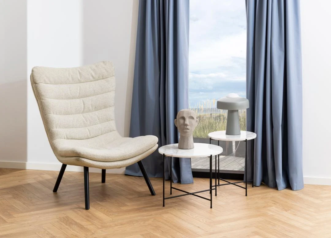 Een moderne kamer met een beige fauteuil, twee Bijzettafel Zwart, Wit 52 CM Rond kunststof bijzettafels, een gebeeldhouwd hoofd, grijze lamp en blauwe gordijnen die een groot raam met uitzicht naar buiten omlijsten.
