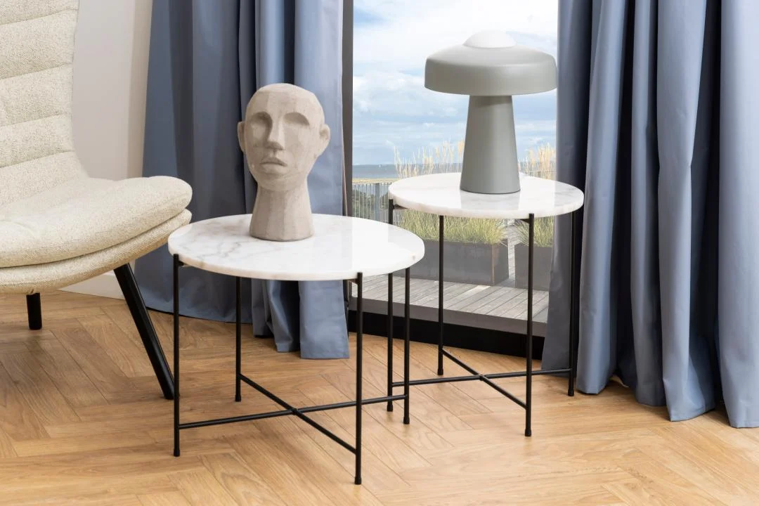 Twee Bijzettafels Zwart, Wit 52 CM Rond kunststof staan bij een raam met blauwe gordijnen. Op de ene staat een grijs gebeeldhouwd hoofd, op de andere een moderne grijze lamp. Een beige stoel is gedeeltelijk zichtbaar aan de linkerkant.