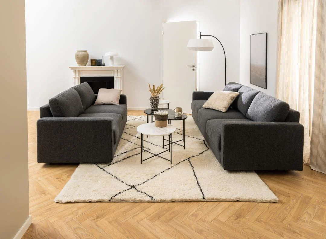 Twee donkergrijze banken staan tegenover elkaar op een licht vloerkleed over een visgraatvloer. In het midden staat de Bijzettafel Zwart, Wit 52 CM Rond, een ronde wit met zwarte kunststof bijzettafel met decor. Gordijnen filteren het licht; een lamp en open haard staan erachter.