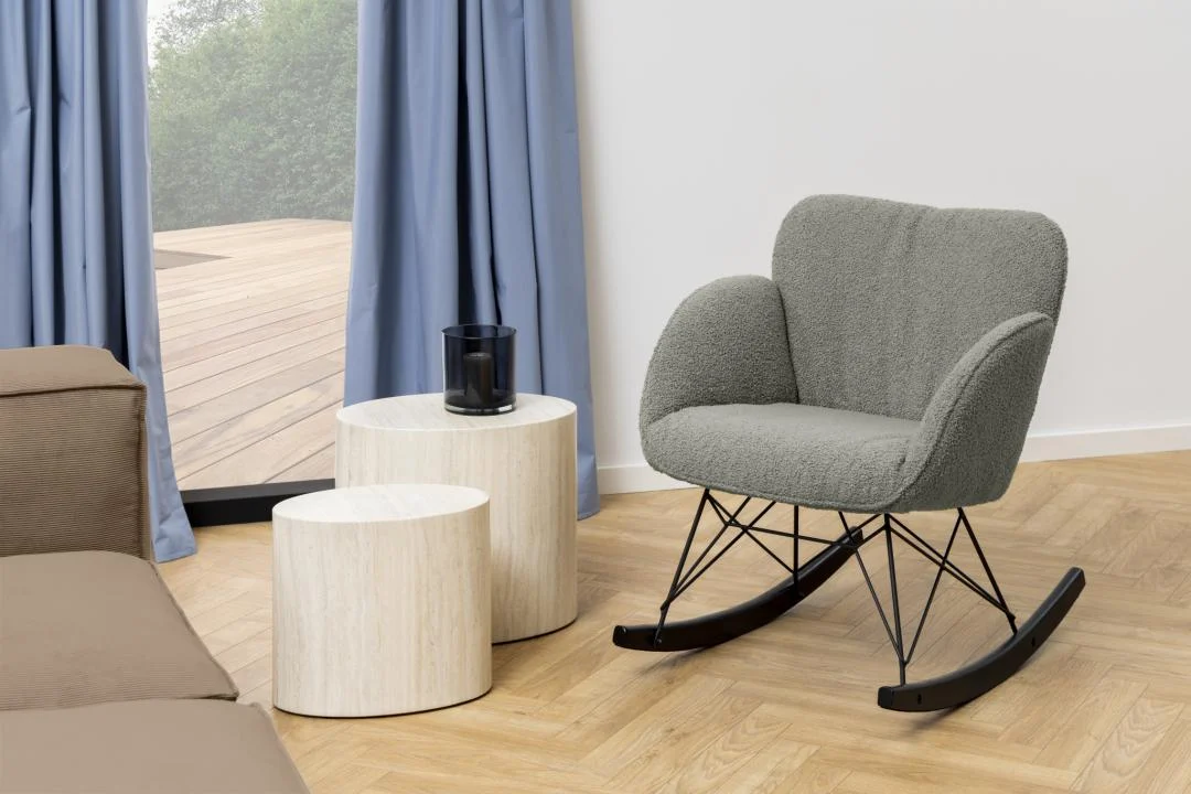 Een moderne woonkamer met een grijs gestoffeerde schommelstoel, twee ovale beige Salontafels 33x40 cm van kunststof, een kandelaar van zwart glas, een lichthouten vloer en blauwe gordijnen bij een glazen deur naar een houten terras.