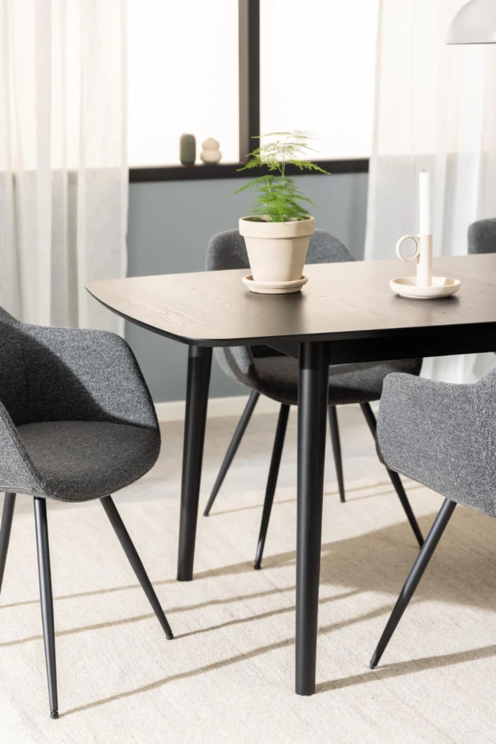 Een moderne eethoek met de Eettafel Montreux 90 x 75 CM Rond MDF Mat met een zwart onderstel, drie grijs gestoffeerde stoelen, een plant in pot en een witte kandelaar. Gordijnen bedekken de hoge ramen op de achtergrond.