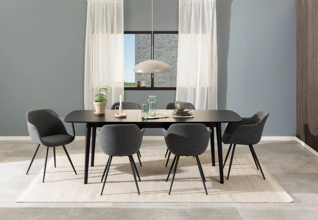 Een moderne eetkamer met de Eettafel Montreux 90 x 75 CM rond MDF mat met een zwart onderstel, omringd door zes donkergrijs gestoffeerde stoelen, een beige vloerkleed, hanglamp, tafeldecoratie, planten, transparante witte gordijnen en een raam op de achtergrond.