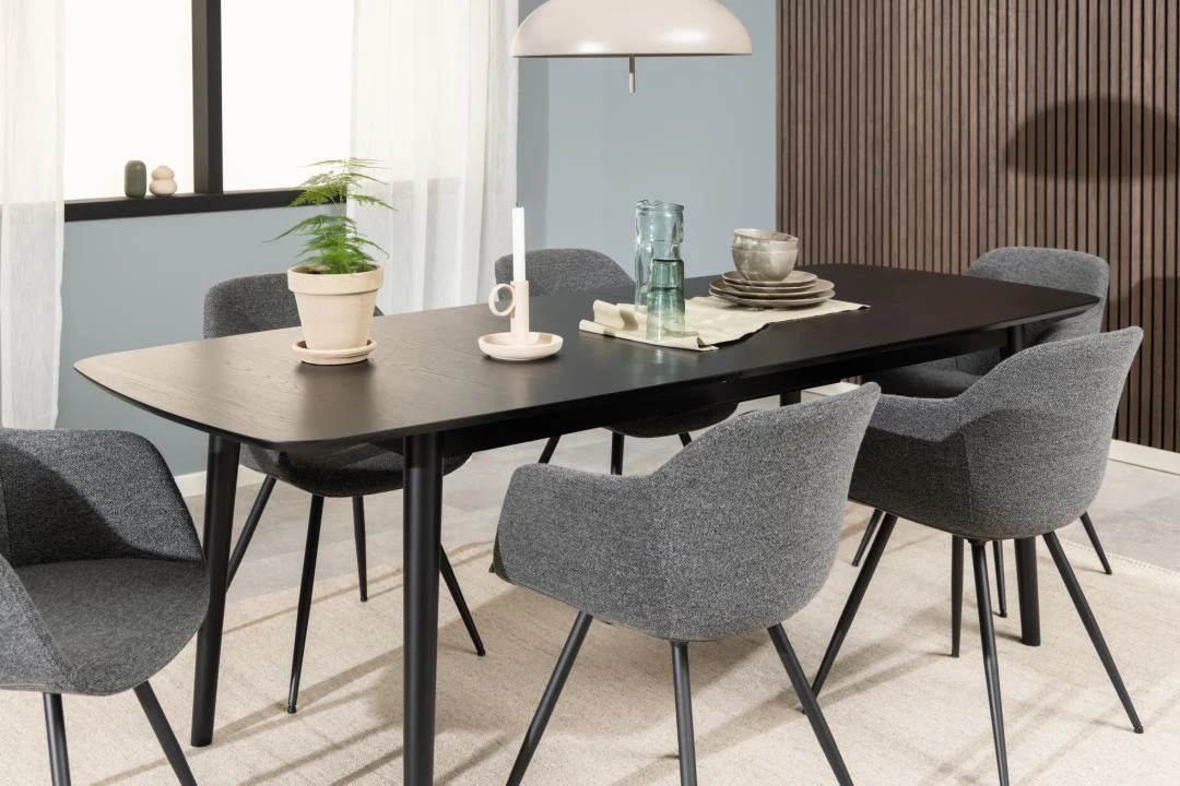 Moderne eetkamer met de Eettafel Montreux 90 x 75 CM ronde MDF mat tafel met zwart onderstel, zes grijs gestoffeerde stoelen, een potplant, servies, glaswerk, een kaars en een hanglamp tegen een blauwe muur en een wit venster.