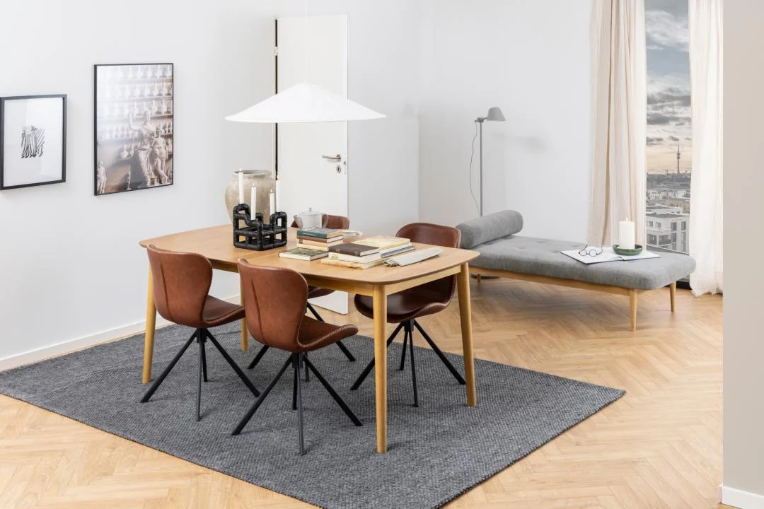 Een moderne eetkamer met de Eettafel Nieuwe Sfeer 219 x 90 CM Rond gemaakt van eikenhout, vier bruine stoelen, boeken en decor op tafel, een grijs vloerkleed, een grijs daybed bij grote ramen met uitzicht op de stad, minimalistische muurkunst en een staande lamp.