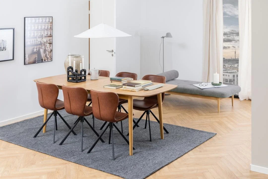 Een moderne eetkamer met de Eettafel Nieuwe Sfeer 219 x 90 CM Rond in eikenhout, zes bruine stoelen, boeken en decoraccenten, grijs vloerkleed, muurkunst, dagbed, hoge lamp, groot raam met uitzicht op de stad, lichte houten vloeren en witte muren.