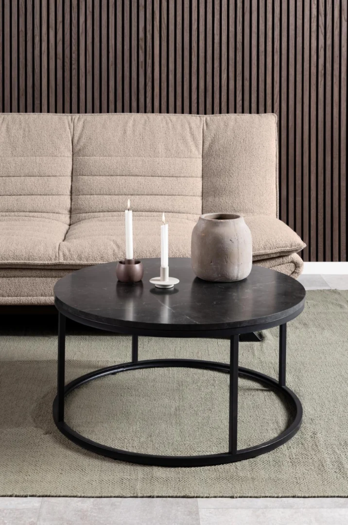 Een moderne woonkamer met een beige bank en een Salontafel Zwart 80 CM Rond Kunststof (industrieel), met twee witte kaarsen en een vaas van keramiek, op een groen vloerkleed met daarachter een accentmuur van houten latten.