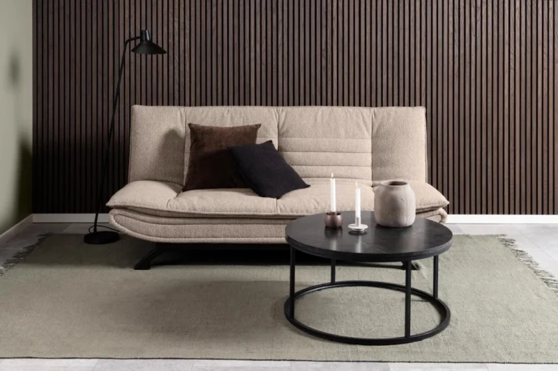 Een moderne woonkamer met een beige bank, twee donkere kussens, een zwarte staande lamp, een licht vloerkleed, verticale bruinhouten wandpanelen en een industriële Salontafel Zwart 80 CM Rond Kunststof met kaarsen en een vaas.