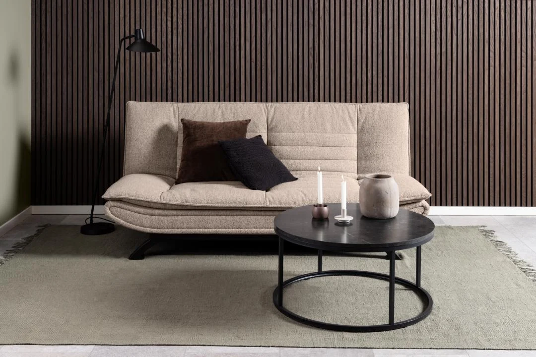 Een moderne woonkamer met een beige bank, twee donkere kussens, een zwarte staande lamp, een licht vloerkleed, verticale bruinhouten wandpanelen en een industriële Salontafel Zwart 80 CM Rond Kunststof met kaarsen en een vaas.
