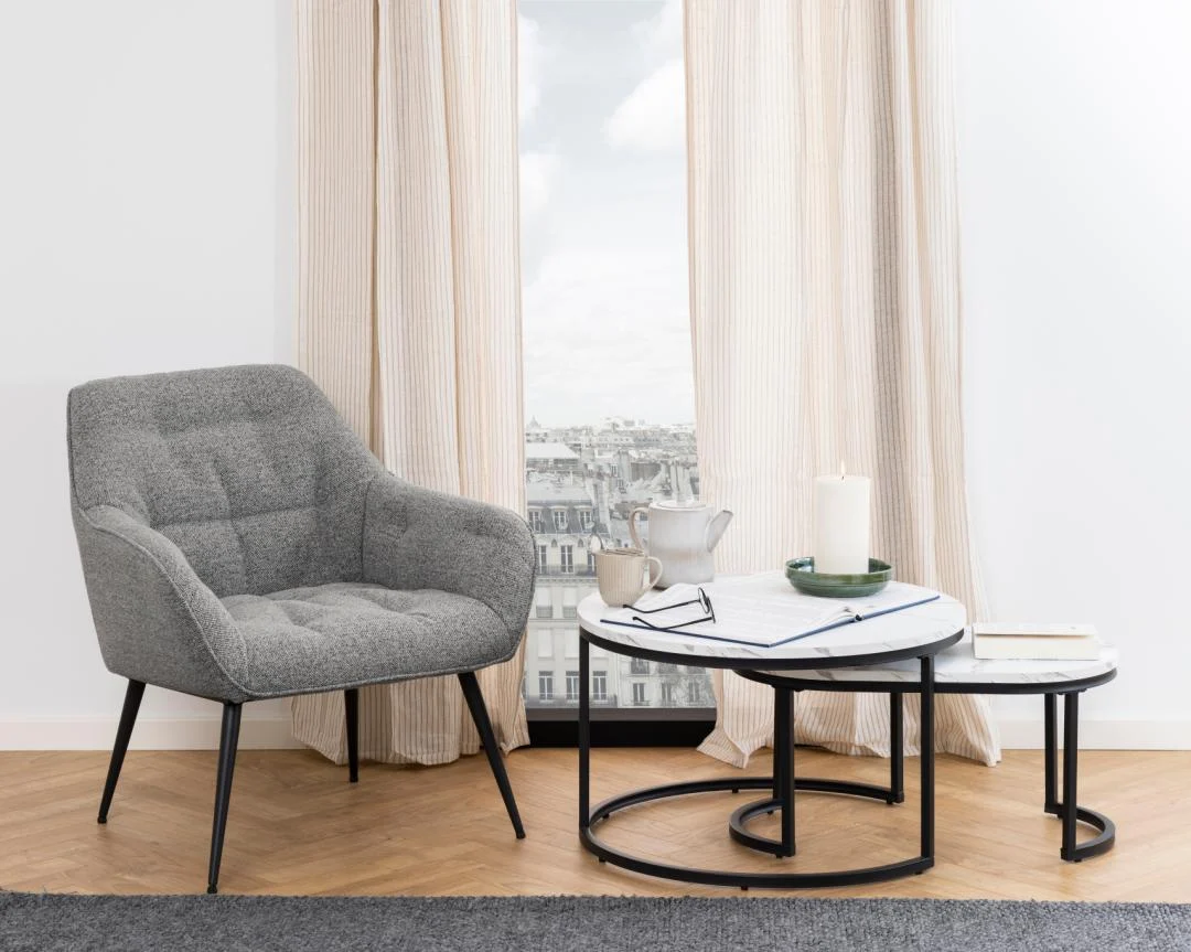 Een moderne woonkamer met een grijze fauteuil, een Salontafel Zwart, Wit 70 x 42 CM Rond van kunststof, een kaars, theepot, boeken en kopjes. Lichte gordijnen zijn open om een stadsgezicht door het raam te onthullen.