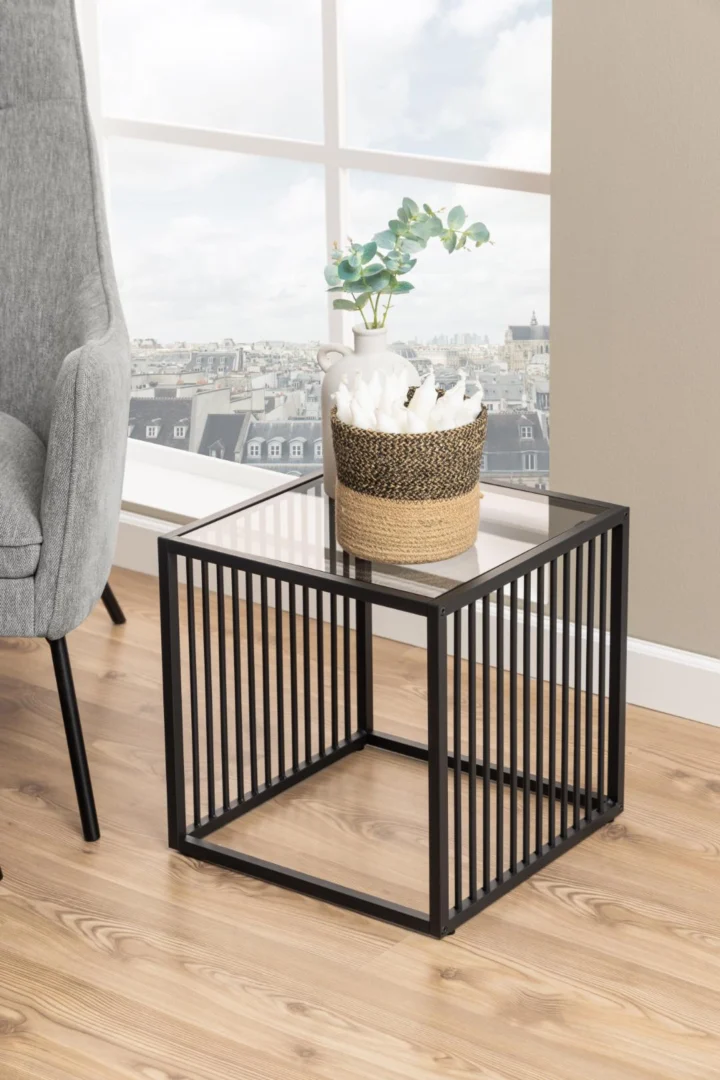 De Bijzettafel Grijs, Roest (40 x 40 cm) met een vierkant stalen frame staat op een houten vloer. Daarop staat een gevlochten mand met een plant en een wit decor, naast een grijs gestoffeerde stoel bij een groot raam met uitzicht op de stad.