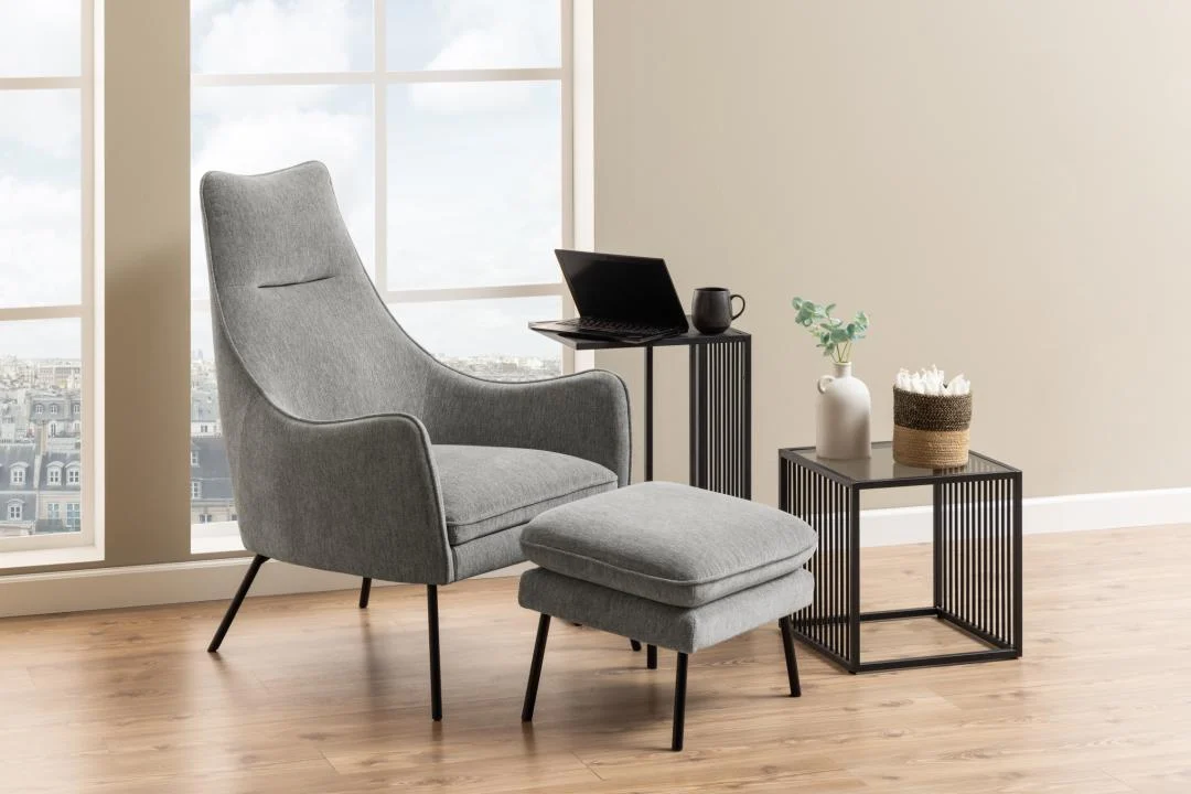 Een moderne woonkamerhoek met een grijze fauteuil, voetenbank en de Bijzettafel Grijs, Roest 40 x 40 CM-een vierkante stalen bijzettafel met een laptop en een plant in pot. Grote ramen en een houten vloer maken de gezellige opstelling compleet.