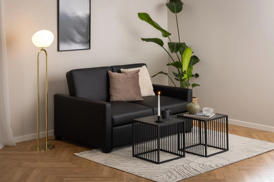 Een moderne woonkamer heeft een zwarte bank met beige en bruine kussens, twee vierkante stalen bijzettafels van Bijzettafel Grijs, Roest 40 x 40 CM, een brandende kaars, plant in pot, staande lamp met ronde kap en een ingelijst abstract kunstwerk aan de muur.